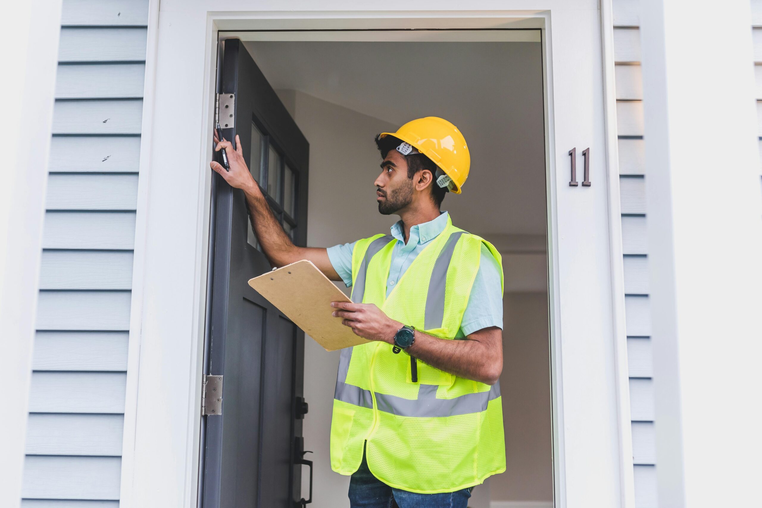 A home inspector in safety vest and hard hat checks doorframe alignment with clipboard outdoors.